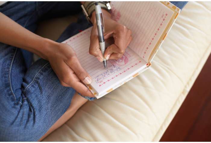 How to be More Productive in 2022 Tip 5: Reflect and journal to document lessons learned and ideas to make things better. Photo features a woman sitting on a white puffy blanket, journaling in a small notebook.