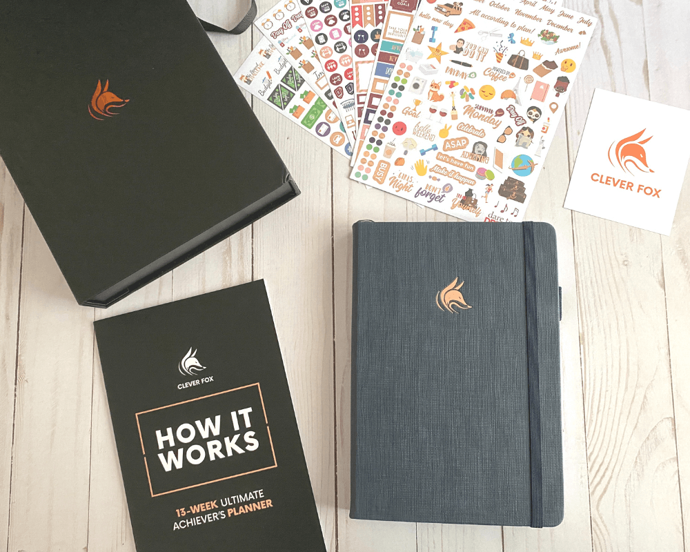 desk flatlay of planner and various accessories on a light wood background. top left shows a black keepsake box with a bronzish fox emblem, middle top includes 6 pages of colorful planner stickers, to the right a small card with the clever fox logo and company name, bottom left is a black and white "how it works" guide and bottom right is a navy/dark gray Clever Fox Ultimate Achiever's 13 week planner with a bronzish fox emblem on the hardcover with a dark gray elastic and pen loop. all items shown come with purchase of the Ultimate Achiever's 13 week planner.